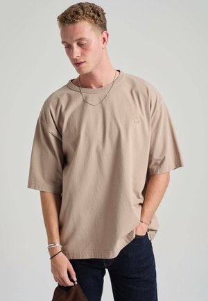 Jeune homme aux cheveux bouclés portant un t-shirt beige oversize, un jean foncé et des bijoux en argent, regardant vers le bas avec une main dans la poche.