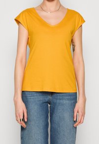 Gul bomull t-shirt med V-ringning, korta uppvikta ärmar och en avslappnad passform, kombinerad med blå högmidjade jeans.