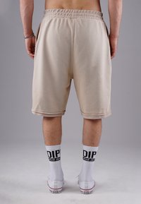Beige athletic shorts with elastic waistband. Smooth texture, knee-length, paired with white socks featuring black lettering and casual sneakers.