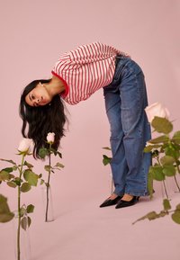 Naine triibulise särgi ja teksadega kummardumas ettepoole, haistmas klaasvazodes heleroosasid roose pehme roosa tausta vastu.