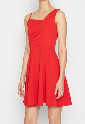 Femme portant une robe rouge sans manches avec un col asymétrique et une jupe évasée, debout devant un fond clair uni.