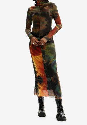 Femme portant une robe multicolore tie-dye à manches longues et col haut, associée à des bottines noires lacées, debout les mains jointes.