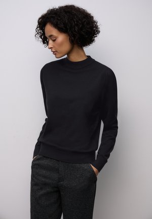 Woman with curly black hair wears a long-sleeve black sweater and dark gray textured pants, standing with hands in pockets, looking down left.