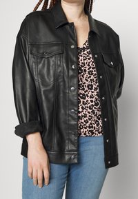 Veste en cuir noir à boutons avec deux poches poitrine et des accessoires en métal argenté, superposée à un top à imprimé léopard rose, associée à un jean bleu.