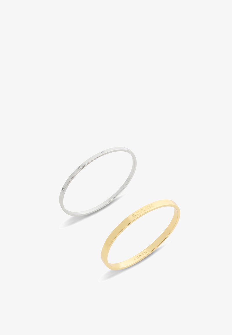 Par de pulseras: una de plata con pequeños cristales transparentes, una de oro con el nombre de la marca grabado. Ambas tienen un diseño delicado y suave.