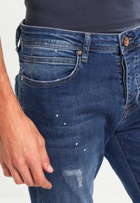 Vyšúchané modré denimové džínsy so škvrnami od farby, opotrebovanými detailmi, s gombíkovým zapínaním a viacerými vreckami.