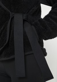 Zwart, pluizig stoffen wikkel met een gestructureerd oppervlak, met een brede zwarte strik in de taille en gladde accenten op de mouwen.