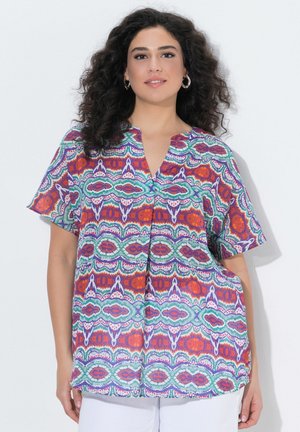 Vrouw met donker krullend haar, draagster van oorringen en een kleurrijk patroon blouse met korte mouwen, staand tegen een witte achtergrond.