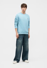 Νέος άνδρας στέκεται φορώντας ανοιχτό μπλε φούτερ, φαρδιά σκούρα μπλε τζιν και λευκά αθλητικά παπούτσια μπροστά σε απλό γκρι φόντο.