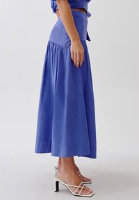 Una lunga gonna fluida in cotone blu solido, con un alto punto vita e design arricciato, abbinata a sandali bianchi con cinturini.