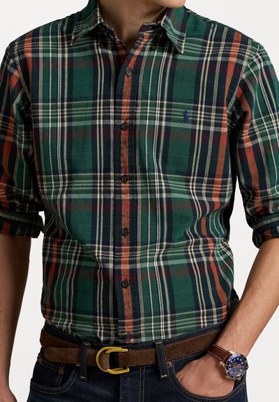 Camisa de cuadros verde abotonada con acentos en naranja y azul marino, de material de algodón, de manga larga con los puños remangados, combinada con un cinturón y un reloj marrones.