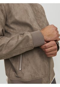 Chaqueta bomber de ante marrón con cremallera plateada y puños acanalados, con una textura suave y un diseño aerodinámico.
