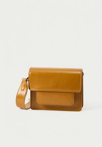 Καφέ δερμάτινη τσάντα χιαστί με εμπρός καπάκι και φαρδιά ρυθμιζόμενη λουρί ώμου πάνω σε απλό ανοιχτό φόντο.