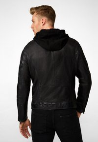 Zwarte leren jas met een gestructureerde afwerking en zichtbare naden, voorzien van een zwarte capuchon. Gecombineerd met donkere jeans, wat een casual ontwerp laat zien.