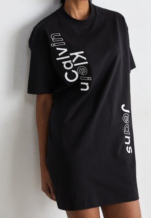 Person iført en sort, overdimensioneret t-shirt-kjole med hvid tekst "Calvin Klein" diagonalt og "Jeans" lodret på forsiden.