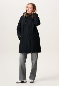 Vrouw in een zwarte gewatteerde jas met capuchon, grijze jeans en witte sneakers, staande met één hand in het haar en de andere in de zak van haar jas.