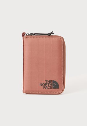 Portafoglio compatto quadrato rosa cipria con zip nera e logo The North Face sul davanti su sfondo uniforme.