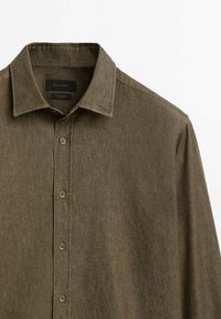 Chemise à manches longues vert olive avec boutons, col classique et coutures visibles sur les épaules et la patte de boutonnage avant.