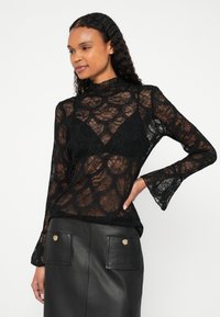 Mustast pitsist pluus kõrge kaeluse ja lehvivate varrukatega, läbipaistva tekstuuri ja geomeetrilise mustriga, kantud musta rinnahoidja peale.