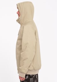 Beige hættetrøje med lynlås foran, elastiske manchetter og kant. Glat stoftekstur med et enkelt design; minimale accenter og ingen mønstre.