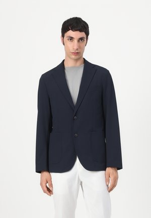 Ung mand iført en mørk navy blazer over en grå skjorte og hvide bukser, stående mod en ensfarvet hvid baggrund.