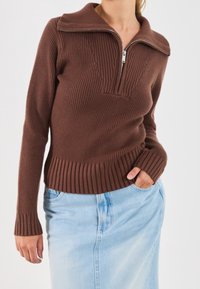 Pull marron côtelé avec un col à fermeture éclair et un design en forme de V, associé à une jupe en denim clair. Tissu texturé et manches ajustées.