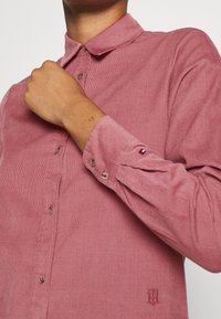 Roze ribfluwelen shirt met een button-down kraag, lange mouwen en gestikt logo-detail op de borst. Kenmerkt zich door een verticale ribbeltextuur.