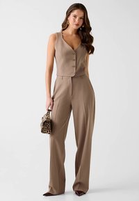 Gilet beige sur mesure avec un col en V et des boutons, associé à un pantalon à jambes larges. La tenue présente une finition texturée et un petit sac à main imprimé léopard.