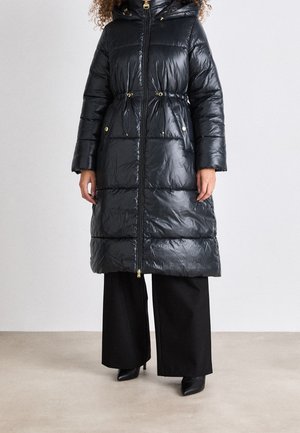 Woman wearing a long, shiny black puffer coat with drawstrings at the waist, paired with wide-leg black pants and black pointed boots.