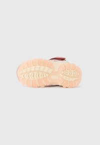 La semelle extérieure de la chaussure de sport présente une semelle intermédiaire en caoutchouc beige rugueux et un motif de grip texturé avec des rainures profondes. Accentuée de détails rouges.