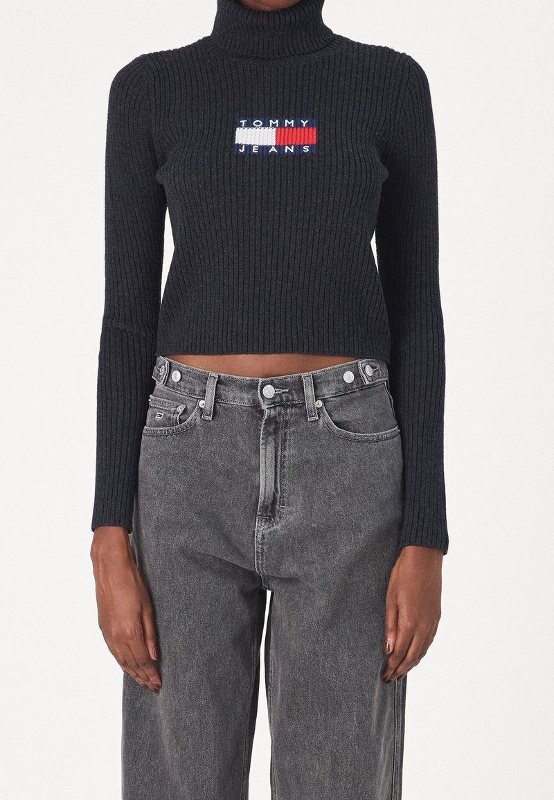 Pull court noir côtelé à col roulé avec logo Tommy Jeans et jean noir délavé taille haute porté par une personne sur un fond uni.