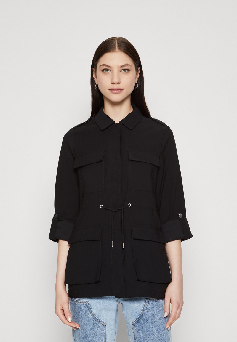 Giacca utility nera con collo a camicia, vita regolabile con cordino e due tasche frontali. Realizzata in tessuto leggero con maniche arrotolate.
