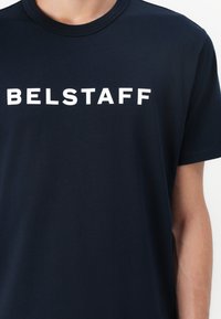 Jūras zilo kokvilnas t-kreklu ar vārdu "BELSTAFF" treknos baltajos burtiem uz krūšu daļas. Klasiska apaļa apkakle un īsās piedurknes.