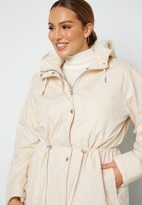 Béžový kabát se zipem vpředu, stažení v pase na šňůrku a kapucí. Má stříbrné kování a měkkou podšívku. Kapsy na obou stranách.