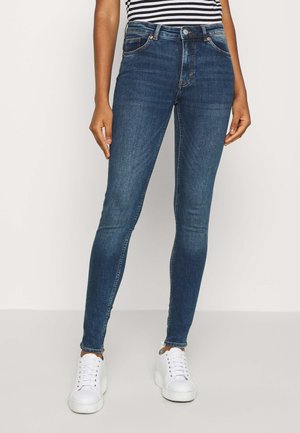 Jeans de mezclilla azul ajustados con una cintura de altura media, que cuentan con un diseño clásico de cinco bolsillos y un ligero desgastado. Combinados con zapatillas blancas.