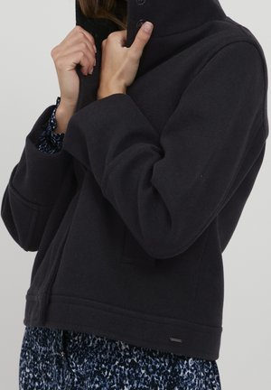 Woman adjusting collar of black wool jacket over blue and white patterned dress against plain background.