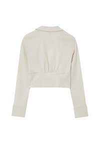 Veste beige raccourcie avec des manches longues, un col plat et un dos froncé. Texture lisse avec une taille cintrée et des poignets larges.
