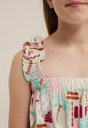 Jeune fille portant une robe couleur crème avec des motifs floraux multicolores et abstraits et une bretelle à volants.