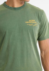 Olivgrünes Baumwoll-T-Shirt mit Rundhalsausschnitt und kurzen Ärmeln, versehen mit gelbem "GUESS ORIGINALS"-Text und grafischem Detail auf der Vorderseite.