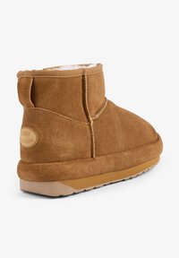 Botas de tobillo de ante marrón con un suave forro blanco, detalles de costura texturizados y una gruesa suela de goma beige con un logo en la parte trasera.