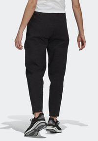 Persoon in zwarte tapered broeken en zwarte Adidas sneakers, die op een lichtgrijze ondergrond loopt, gezien vanuit de rug.