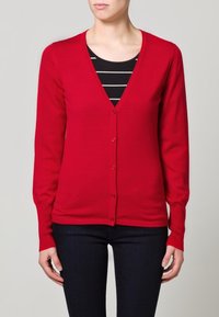 Cardigan rosso realizzato in tessuto morbido, dotato di scollatura a V, maniche lunghe e bottoni frontali. Indossato sopra una maglietta a righe bianche e nere.