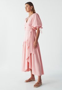 Pink wrap dress with puff sleeves, a deep V-neck, and ruffled hem. Made of soft fabric, featuring ties at the waist. Worn with flat sandals.