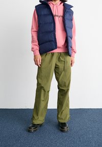 Person som bär en marinblå pufferväst över en rosa hoodie, olivgröna vida byxor och svarta snörstövlar som står på en blå matta.