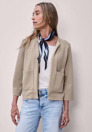 Femme portant une veste beige ouverte avec de grandes poches frontales, un haut blanc, un jean bleu clair et un foulard noir et blanc rayé autour du cou, regardant vers la gauche.