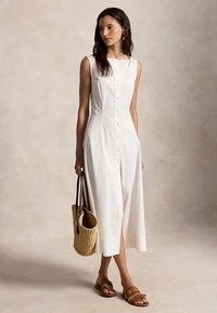Woman in sleeveless white button-front midi dress holding straw tote bag, wearing brown flat sandals, standing against plain background.