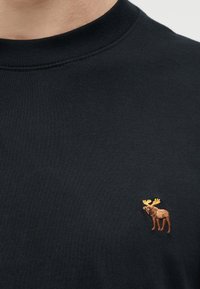 Κοντινό πλάνο μαύρου μπλουζιού με στρογγυλή λαιμόκοψη και μικρή κεντημένη  καφέ άλκη με κίτρινα κέρατα στο στήθος.