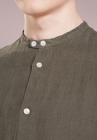 Olivově zelená lněná košile s kulatým výstřihem, třemi bílými knoflíky na zapínání a texturovaným detailem.