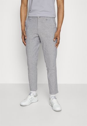 Mężczyzna ubrany w jasnoszare spodnie typu slim fit, białe trampki z szarymi detalami oraz jasnoszarą koszulkę, stojący na białym tle.
