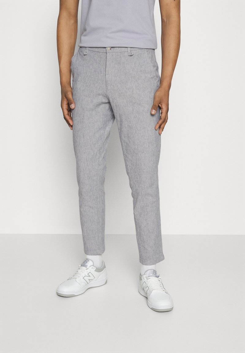 Homme portant un pantalon slim gris clair, des baskets blanches avec des détails gris et un t-shirt gris clair, debout devant un fond blanc.